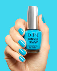 Hand with glossy teal nails holds an OPI Infinite Shine Beach Party Teal Dawn gel-like lacquer bottle against a bright turquoise background.