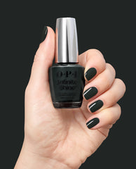 Hand holding OPI Infinite Shine Nein! Nein! Nein! OK Fine! nail lacquer, with glossy deep forest green nails showcasing a bold shade for fall nail colors against a dark background.