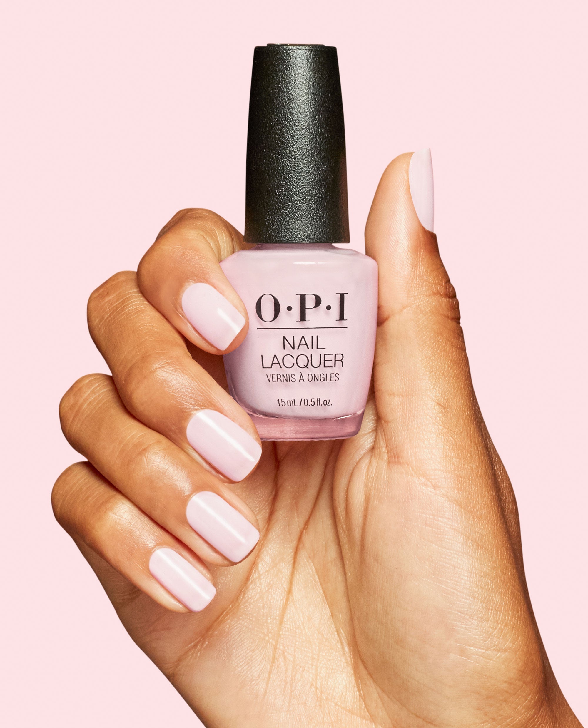Hand with glossy pale pink manicured nails holds an OPI Nail Lacquer bottle in OPI’m a Bubble Bunny, a soft pastel pink shade, against a light pink background.