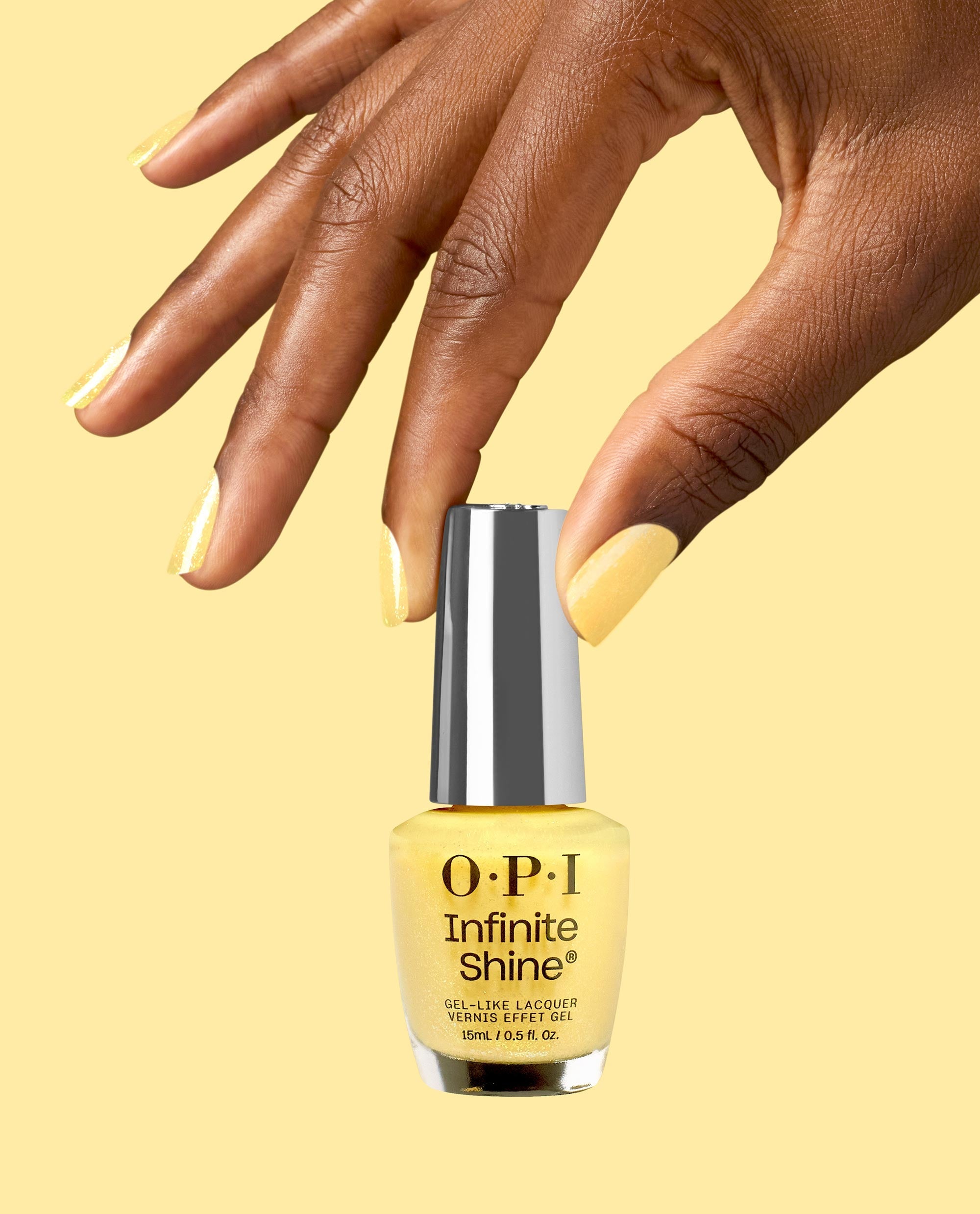Hand with glossy yellow nails holds an OPI Infinite Shine Slay Hello to Yellow nail polish bottle, a soft shimmer yellow gel-like lacquer, against a pale yellow background.